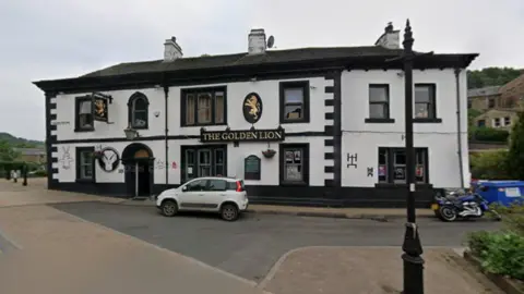 Google The exterior of a large building set on the side of street. The building is painted in white and black. On the front is a sign in gold letters which reads 'The Golden Lion'.