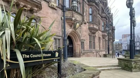 The Council House in Coventry, a large brown/red stone bricked building with arches and buttress windows. In front of the arched opening is a paved area with a couple of steps and a raised stone flower bed with some long-leaved green bushes slightly covering a sign which says "The Council House".