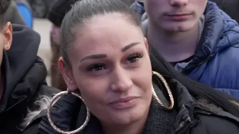 BBC Victoria Phillips has scraped back hair and big hooped earrings. She is looking away from the camera and giving an interview. There are people behind her. 