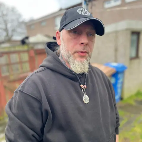 Ronnie McLellan looking straight at the camera. He is wearing a black hat and a black hoodie. He has a grey beard and a silver chain round his neck.
