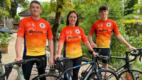 Gavin Kermack / BBC The same woman back in the garden, with a middle-aged man and a teenage boy, all wearing the same lycra tops. They are smiling at the camera and all are resting their hands on bicycles.