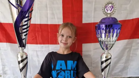 Isla, a 12-year-old dancer, has her blonde hair back in a ponytail and is wearing a black t shirt with 'I am a world champion' written on it. She is holding two bit trophies, which are silver, black and purple in colour. She is standing in front of an England flag and smiling at the camera.