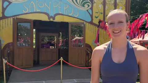 Chelsea stands in front of the Norfolk and Norwich Festival's yellow circus tent on a sunny day. She is smiling, wearing a purple halterneck leotard. Her hair is blonde and pulled back from her face.