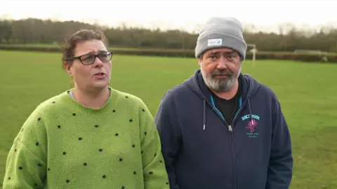 A woman wearing a green jumper with black dots and a man wearing a grey jacket and a light grey beanie hat stand in a field. A grass lawn can be seen in the background behind them.