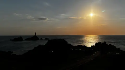 The silhouette of the coastline and lighthouse on the rocks at Corbiere in Jersey with a small circle of golden sunlight glows in the sky above and reflects on the sea amid dusky blue skies.