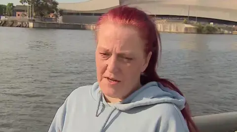 Rebecca Ramsay with long auburn hair in a pony tail wearing a pale blue hoodie stands in front of Salford Quays. She is frowning as she describes her heartache at the tragic deaths of teenagers in open water.