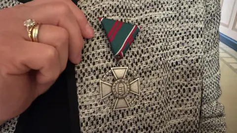 A close up of a silver medal pinned to a woman's jacket. It is a silver cross with a green and red fabric attached to it.