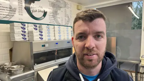 Miguel Costa has short brown hair and matching beard and moustache, he has a dark blue hoodie on with a sky blue shirt underneath, behind him is equipment in a fish and chip shop, and a price list board is above on the wall to the left.