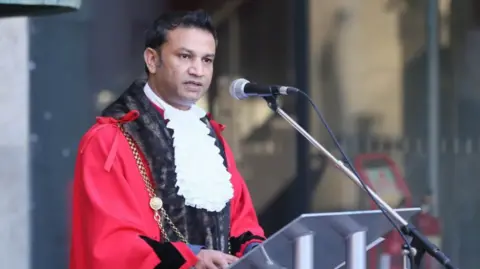 Newcastle City Council A man in a red robe with brown fur trim and white ruffled shirt standing in front of a microphone, giving a speech.