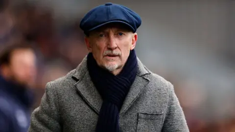 Swindon Town bos Ian Holloway wearing large grey overcoat, dark blue woolen scarf and royal blue cloth flat cap