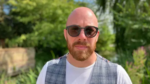 Scott Johnson standing outside with a garden, wall and trees behind him. He is wearing dark glasses and has a beard. He is wearing a grey plaid waistcoat over a white T-shirt.