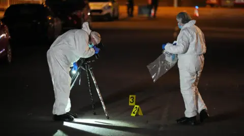 Two crime scene investigators, dressed in hazmat suits, is conducting an investigation on a road. One of them is taking photographs while the other one is holding a bag to store crime scene evidence. 