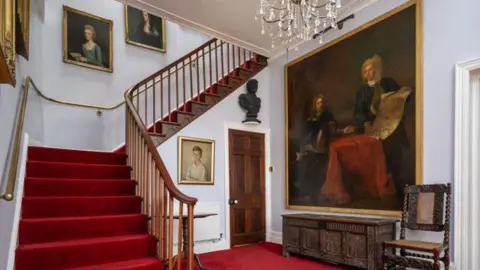The Armstrong Family The picture shows a grand entrance hall inside a historic house, dominated by a sweeping staircase covered in deep red carpet. The staircase curves gently upwards to the left, with polished wooden banisters and spindles, and continues to a landing above. On the right-hand wall, there is a very large oil painting in a gilt frame showing two seated figures.