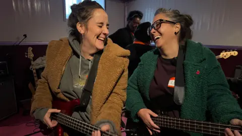 Michelle and Lou hold guitars in the rehearsal studios and laugh together. 