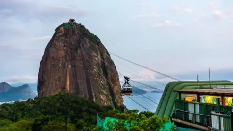 Emin Sansar/Anadolu via Getty Images A view of Sugar Loaf cable car at Guanabara Bay in November 2024. The steep rocky mountain can be seen against the blue of the bay. At the top of the mountain, the cable car station is visible. 
Cables connect it with Morro da Urca, from which the photo is taken. A cable car can be seen suspended halfway between the two. 