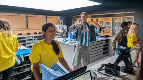 A smiling woman with a yellow tshirt standing behind a low desk with rows of shelves behind her and other people in the room