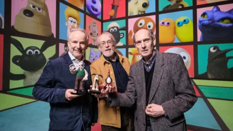 Aardman animation's co-founder and creators are standing in front of screen showing a collection of the studios most famous characters, including Wallace & Gromit, Shaun the Sheep and Morph. Peter Lord is standing in the middle wearing a brown Jacket holding a Gromit figurine. Nick Park is standing to the left of him wearing a navy blazer and holding a Wallace figurine and David Sproxton is standing to the right of Peter holding a penguin figurine. 