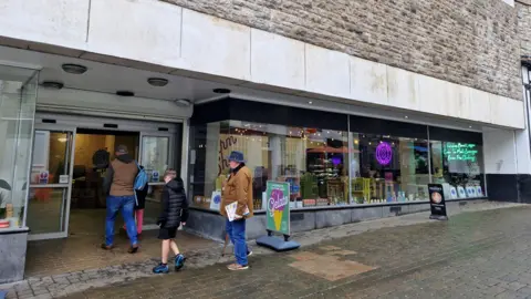 The front of a shop with various illuminated signs in the window. Two men wearing brown jackets and blue jeans, and a boy in shorts and a black coat are walking into the open double doors. There is a sign outside advertising Gelato ice cream.