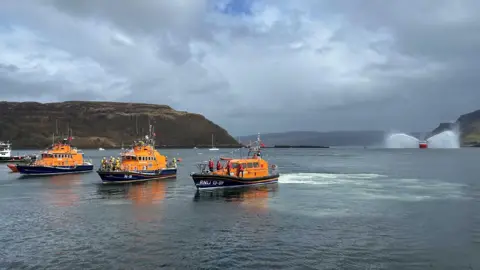 Three orange and blue coloured lifeboats can be seen travelling through a narrow bay. Behind them in the distance is a larger coastguard ship spraying its water cannons. 