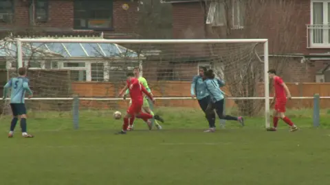 A goal-mouth scramble involving a team of players in red kits and a team in blue. A defender for the red team kicks the below away from the goal in front of the goalkeeper as players in blue rush towards them. They are playing on a grass pitch in front of some red brick homes and a conservatory. 