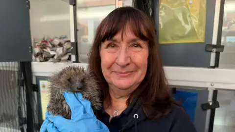 The photo shows a woman looking at the camera. She has brown short hair, and is smiling. She's wearing a navy blue hoodie, and bright blue gloves. She's holding a hedgehog. Behind her are stacks of crates. 