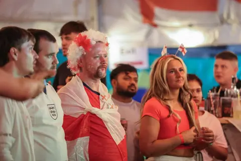 AFP via Getty Images England supporters watch the television broadcast of the 2026 World Cup at a pub in Hebburn, north-east England, with Enland flags draped behind them and wearing England costumes.
