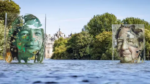 National Lottery/PA An art installation on a park lake. Two faces on clear Perspex screens appear to float on the water.