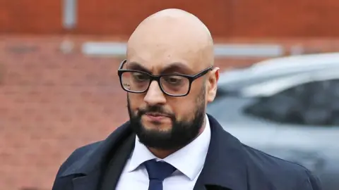 Ben Lack Photography A man with a bald head and wearing glasses, a blue overcoat, white shirt and blue tie looks off camera. Behind him is a brick wall.