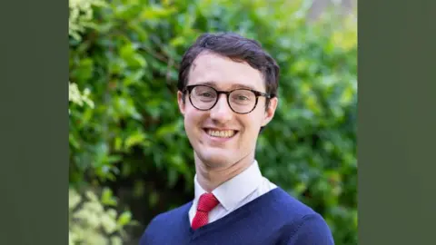 Brian Callingham Dr Eamonn O'Keeffe, who has short dark hair and dark framed glasses. He is wearing a blue v-necked jumper over a white shirt and red tie. He is smiling towards the camera and behind him are the blurred-out green leaves of a tree or shrub.