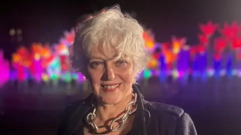 Helen Marriage is standing in front of a bright light installation and smiling at the camera. She has short white hair and is wearing a chunky metal necklace.