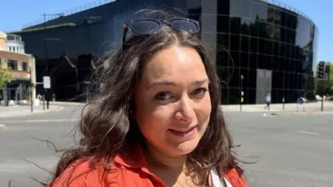 Naomi Drown stands outside the Willis building. She is smiling and has long wavy brown hair. She wears a red shirt with a white T-shirt underneath. Sunglasses rest on top of her head. 