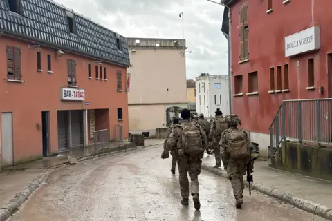 An army exercise is carried out with a group of soldiers running down a muddy road - with a boulangerie sign on the right and a tabac sign on the left. They are carrying backpacks.