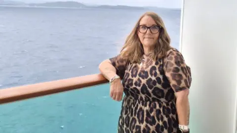 Emma is standing next next to the ocean with her arm rest on a glass ledge. She is wearing glasses and a leopard print dress. 