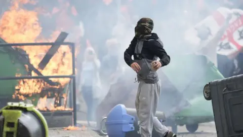 Getty Images A man in a balaclava throws something with protestors and fire in the background. He is surrounded by rubble and turned-over bins