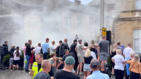 Groups of people throwing bottles and other items at a hotel with smoke in the background