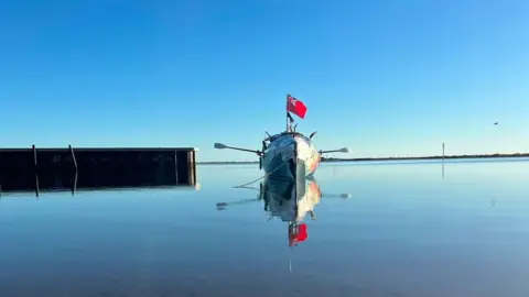 Lara Vafiadis A white row boat with a red flag sticking out from the middle of it. The boat is positioned on calm waters with a pontoon next to it. 