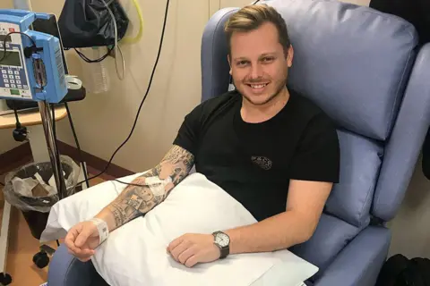 Supplied Ben Uttley, sitting in a cornflower-blue hospital reclining chair and smiling. He is wearing a black t-shirt and has short dark blonde hair, swept back at the front, with a short moustache and beard. He has a black-strapped analogue watch on his left wrist and his right arm - which is covered in tattoos - is resting on a white pillow on his lap. A tube is fed into it and is taped in place. He has a hospital identity bracelet on his right wrist. A piece of hospital equipment, on a metal stand, is on his right.