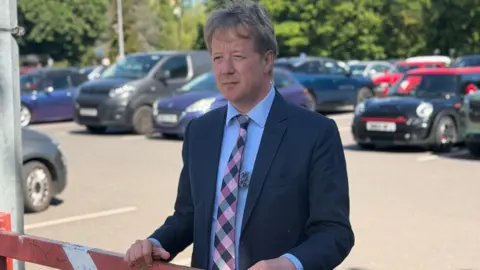 Cambridgeshire and Peterborough Combined Authority A man wears a dark jacket, a blue shirt and a checkered tie. He places his hands on a red barrier at the entrance to a car park.