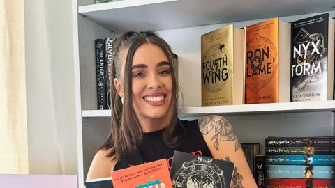 Amelia Lord is a white woman in her late 20s. She has shoulder length brown hair partly pulled back in a ponytail with frontpieces either side of her face. She has defined eyebrows and is wearing makeup, has a central nose ring and earrings, and is smiling at the camera. She wears a sleeveless black top. She is holding a pair of books and stands in front of a bookshelf with collections of books on it, including titles by Rebecca Yarros and the Harry Potter series by JK Rowling. 