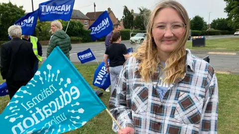 BBC/Seb Cheer A young woman looks at the camera, wearing glasses and a chequered shirt. She is holding a flag with "national education union" written on it, with other people carrying flags reading "NASUWT" in the background.