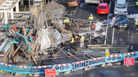 An aerial view of a huge pile of scaffolding poles and wooden planks blocking First Avenue in Hove, which is cordoned off with blue plastic barriers and road closed signs. Workmen in yellow hard hats and high vis jackets are starting to clear the mess. Damaged cars can be seen in the background.