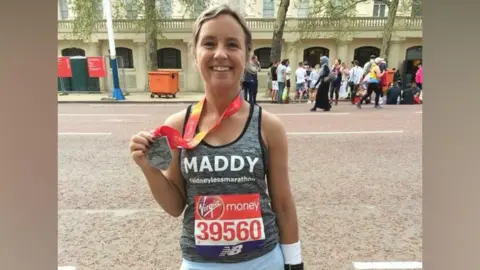 A woman smiles at the camera in a runner's vest with a marathon number stuck to it. She holds up a silver medal around her neck.