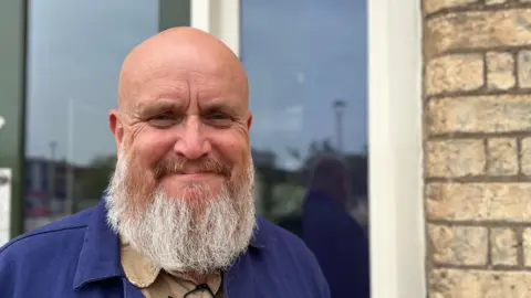 BBC A man with a bald head and a grey beard stands outside in front of a window. He is smiling and wearing a blue jacket.