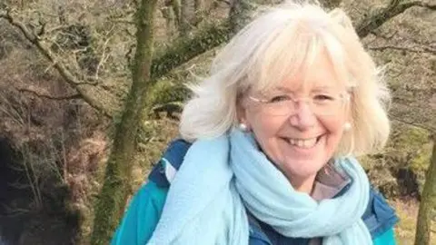 Catherine Sams is standing in a woodland on a sunny day. She is wearing clear-framed glasses, pearl earrings, a blue scarf and a blue raincoat. 