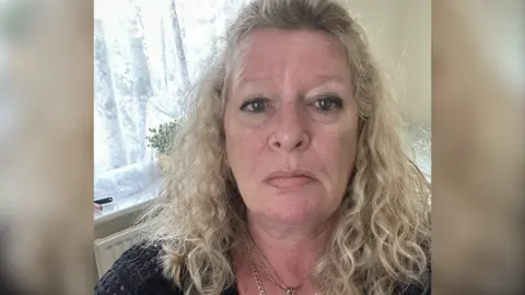 Laraine Chung A head and shoulders selfie-style photo of Laraine Chung. She is pictured in a lounge at home, with a house plant in a window behind her. She has blonde curly hair falling just below her shoulders and is wearing a black crocheted-style top and two gold necklaces. She is looking straight into the camera and is not smiling. 