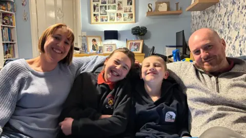 A family of four sit on a sofa in a cosy, busy living room. All are smiling at the camera. On the left is a woman wearing a pale blue jumper. A young girl and a young boy sit next to her with a man on the right wearing a beige jumper.