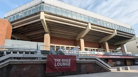 Preston City Council External view of Preston Guild Hall, a large red brick building with steps to the front and a large extension at the top which sticks out and has windows on each side