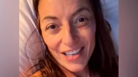 A close up selfie of Davina McCall looking at the camera while lying in a hospital bed, she's smiling as she's talking into camera.