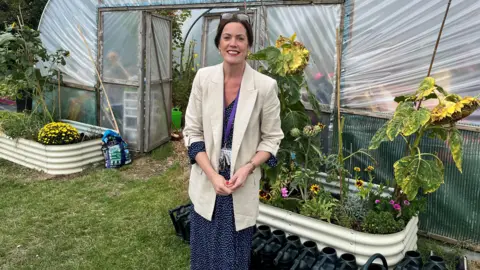 A woman with black hair has a pair of glasses on the top of her head and is wearing a cream jacket and a blue and white spotted dress. She is standing in a garden in front of a greenhouse.