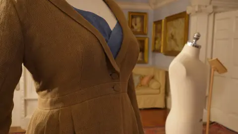 National Trust/Lucy Fenna Close-up of the top of a linen-style v-necked overcoat with two buttons on a mannequin. It is in a room from a stately home with gold-framed pictures on the walls.